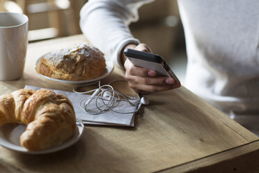 Ausgeschnittenes Bild eines Mannes, der sein Mobiltelefon neben Croissants und Kaffee am Tisch hält - CAVF26180