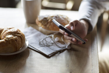 Ausgeschnittenes Bild eines Mannes, der sein Mobiltelefon neben Croissants und Kaffee am Tisch hält - CAVF26181