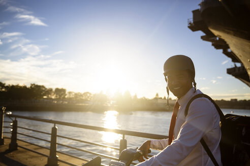 Porträt eines Geschäftsmannes mit Fahrrad, der bei Sonnenuntergang am Fluss steht - CAVF26182