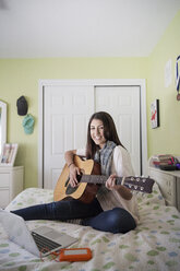 Porträt eines glücklichen Teenagers, der Gitarre spielt und zu Hause am Laptop sitzt - CAVF26194