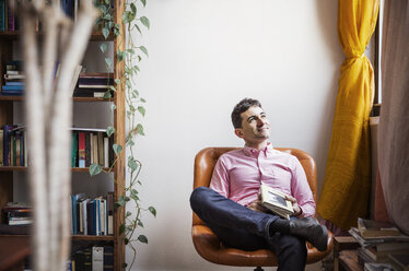 Nachdenklicher Mann hält ein Buch, während er zu Hause auf einem Stuhl sitzt - CAVF26244