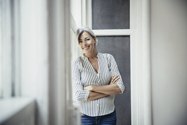 Portrait of happy mature businesswoman standing arms crossed by window in creative office - CAVF26295