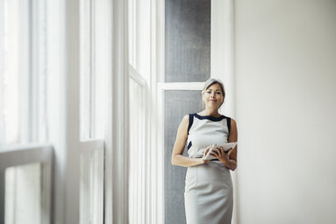 Portrait of confident businesswoman holding tablet computer while standing by window in creative office - CAVF26305