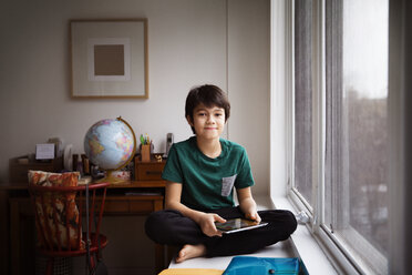 Portrait of boy holding tablet and sitting on table by window at home - CAVF26349