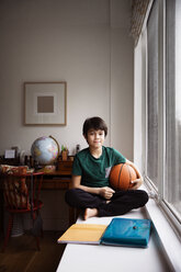Porträt eines Jungen, der einen Ball hält und zu Hause auf einem Tisch am Fenster sitzt - CAVF26350
