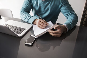 Mittelteil eines Mannes, der an einem Tisch im Büro mit Laptop an einem Buch schreibt - CAVF26361