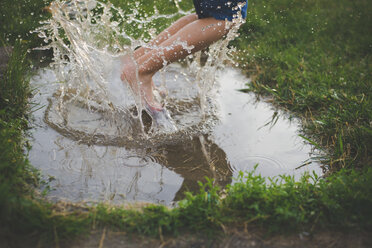 Low section of carefree boy jumping in puddle on grassy field - CAVF26393