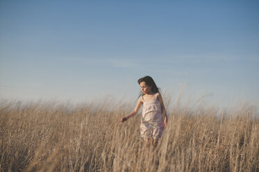 Mädchen berührt trockenes Gras beim Gehen auf einem Feld vor blauem Himmel - CAVF26424