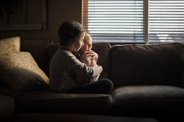 Seitenansicht eines Mädchens, das einen kleinen Jungen umarmt, während es auf dem Sofa zu Hause sitzt - CAVF26470