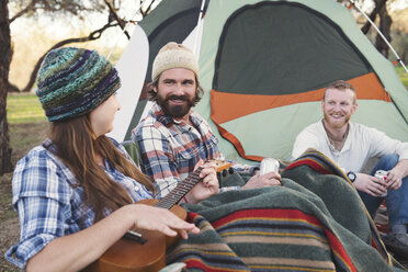 Happy men looking at woman playing guitar outside tent in forest - CAVF26638