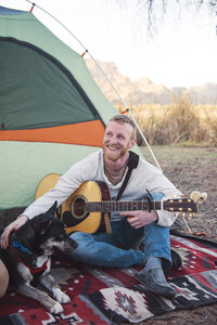 Glücklicher Mann, der seinen Hund streichelt, während er vor dem Zelt eine Gitarre hält - CAVF26641