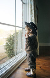 Boy wearing hat while looking through window at home - CAVF26662