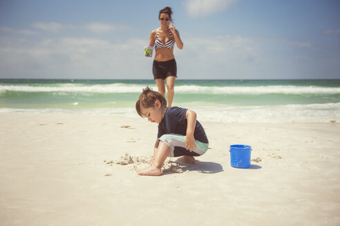 Junge spielt mit Sand, während die Mutter am Strand spazieren geht - CAVF26663