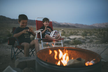 Fröhliche Brüder essen Marshmallows, während sie in der Abenddämmerung auf Stühlen auf dem Campingplatz sitzen - CAVF26675