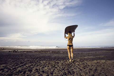 Rückansicht einer Frau, die ein Surfbrett trägt, während sie am Strand gegen den Himmel läuft, lizenzfreies Stockfoto