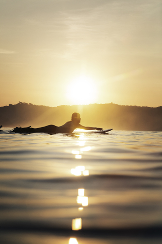 Seitenansicht einer Surferin auf dem Surfbrett liegend im Meer bei Sonnenuntergang, lizenzfreies Stockfoto