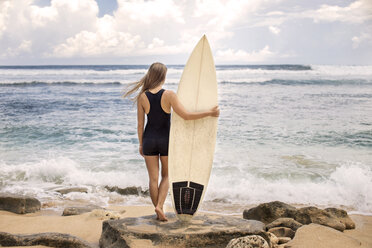 Rückansicht einer Frau, die ein Surfbrett hält, während sie am felsigen Ufer des Strandes steht - CAVF26707