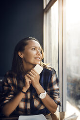 Smiling woman holding smart phone while looking through window at cafe - CAVF26713