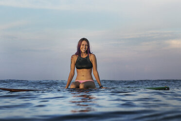 Glückliche Frau sitzt auf einem Surfbrett im Meer bei Sonnenuntergang - CAVF26716