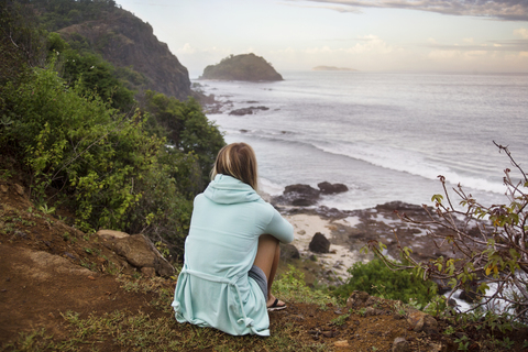 Rückansicht einer Frau, die auf einem Hügel sitzend auf das Meer blickt, lizenzfreies Stockfoto