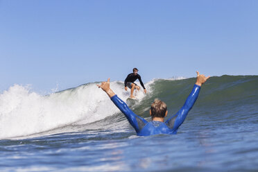 Man cheering while looking at friend surfing on sea against clear blue sky - CAVF26728