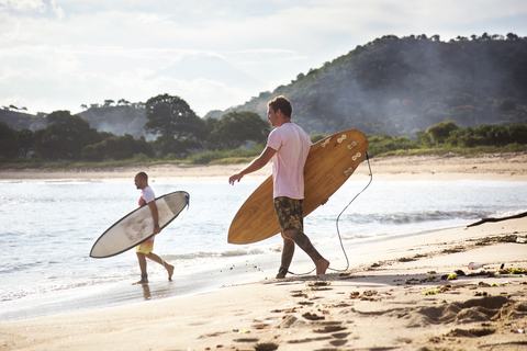Seitenansicht von männlichen Surfern, die Surfbretter tragen, während sie am Strand spazieren gehen, lizenzfreies Stockfoto