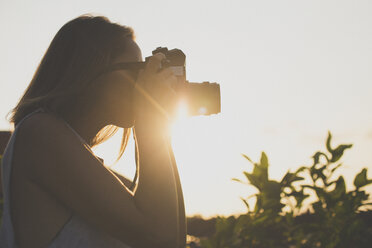 Side view of woman photographing through old-fashioned camera against sky during sunset - CAVF26740