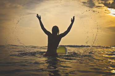 Rückansicht einer verspielten Frau, die auf einem Surfbrett im Meer sitzt und Wasser spritzt - CAVF26769