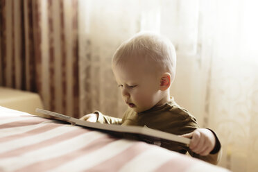 Close-up of boy reading book while standing by bed at home - CAVF26804