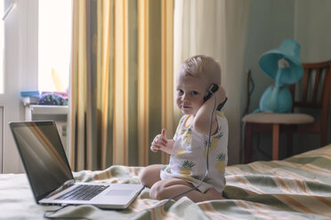 Porträt eines niedlichen kleinen Jungen mit Mobiltelefon und Laptop auf dem Bett zu Hause - CAVF26814