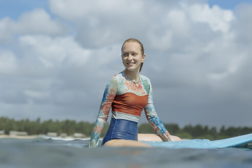 Lächelnde Frau schaut weg, während sie auf einem Surfbrett im Meer gegen den bewölkten Himmel sitzt - CAVF26825
