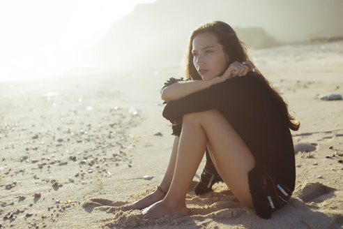 Nachdenkliche Frau, die ihre Knie umarmt, während sie an einem sonnigen Tag am Strand sitzt - CAVF26862