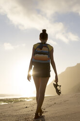 Rear view of woman with backpack holding sandals while walking at beach against cloudy sky - CAVF26866