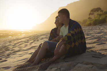 Junges Paar in eine Decke eingewickelt, während es bei Sonnenuntergang den Strand betrachtet - CAVF26868