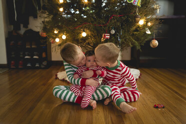 Brüder küssen ihre Schwester, während sie zu Hause am Weihnachtsbaum sitzen - CAVF26920