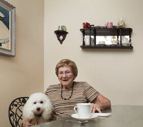 Porträt einer glücklichen älteren Frau, die mit ihrem Hund zu Hause am Tisch sitzt - CAVF26996