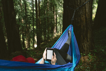 Frau, die einen Tablet-Computer benutzt, während sie in einer Hängematte im Wald liegt - CAVF27277