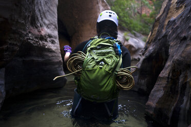 Rear view of hiker with backpack walking in stream amidst rocky mountains - CAVF27296