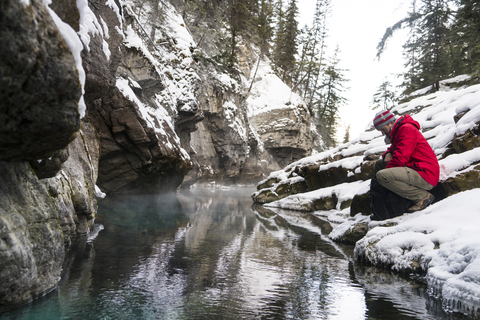 Seitenansicht eines Wanderers mit Rucksack, der im Winter am Bach im Wald kauert, lizenzfreies Stockfoto