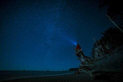 Wanderer mit Stirnlampe auf einem Baumstamm sitzend am Strand von La Push gegen den nächtlichen Himmel - CAVF27348