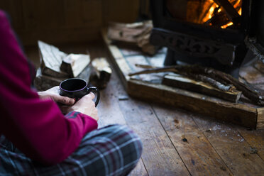 Midsection of man having drink while sitting by wood burning stove in cottage - CAVF27353