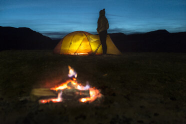 Woman standing by illuminated tent on field against sky during dusk with campfire in foreground - CAVF27381
