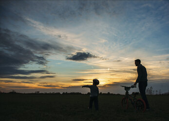 Silhouette von Vater und Sohn mit Fahrrad auf einem Feld bei Sonnenuntergang - CAVF27541