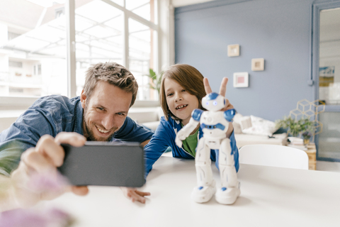 Glücklicher Vater und Sohn machen ein Selfie mit einem Roboter auf dem Tisch zu Hause, lizenzfreies Stockfoto