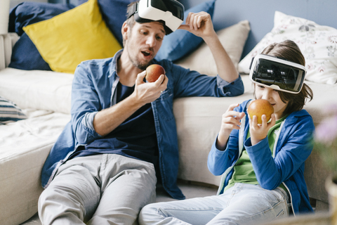 Vater und Sohn tragen VR-Brillen und essen Äpfel zu Hause, lizenzfreies Stockfoto