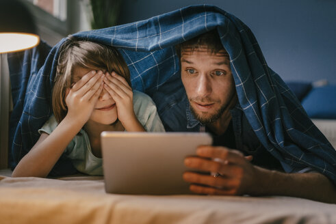 Excited father and son watching a movie on tablet under blanket - KNSF03604