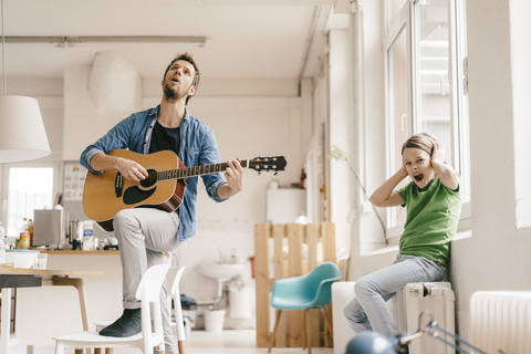 Horrified son covering his ears with father playing guitar at home stock photo