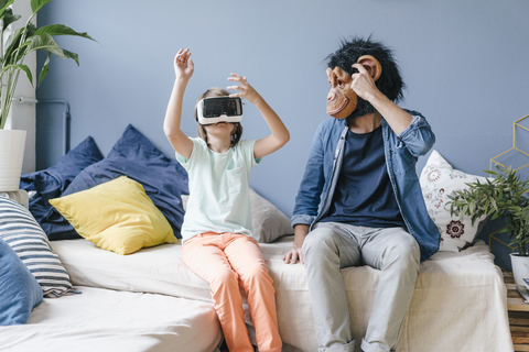 Father wearing monkey mask looking at son wearing VR glasses at home stock photo