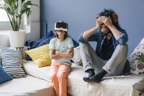Vater mit Affenmaske sitzt zu Hause neben seinem Sohn mit VR-Brille, lizenzfreies Stockfoto