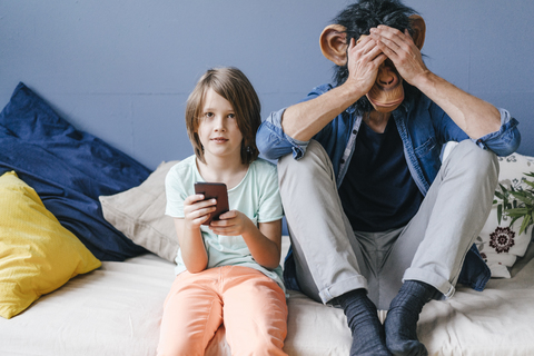 Vater mit Affenmaske sitzt neben seinem Sohn, der zu Hause sein Smartphone benutzt, lizenzfreies Stockfoto